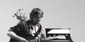 Schwarzweißfoto von Gitarrenlehrer Peter Bachmann mit E-Gitarre vor Klavier sitzend